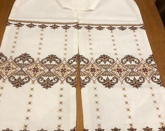 Puede incluir: Un camino de mesa de tela blanca con un patrón simétrico de punto de cruz en tonos marrón y beige. El diseño incluye bandas horizontales de motivos geométricos y florales complejos, complementadas por filas verticales de pequeños acentos en cruz.