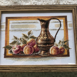 May include: A framed cross-stitch embroidery depicting a still life with a copper pitcher and peaches. The pitcher has an ornate design and is surrounded by peaches and leaves.