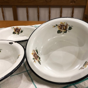 Può includere: Tre ciotole di smalto bianco con bordi neri e disegni floreali marroni. Le ciotole sono su una tovaglia bianca e verde.