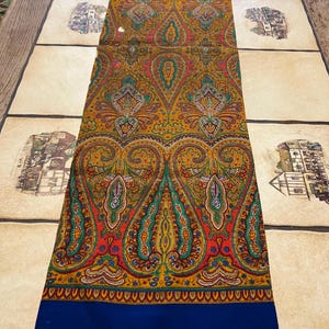 May include: A long, rectangular scarf featuring a colourful paisley design in gold, red, green, and blue. The scarf has a solid blue border at the bottom. The intricate pattern is displayed on a light-coloured surface.