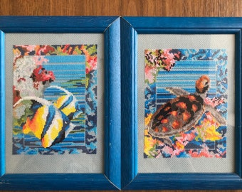 May include: Two framed cross-stitch embroidery pieces. The left piece features a blue and yellow fish swimming in a blue and white striped background. The right piece features a brown and orange turtle swimming in a blue and white striped background.