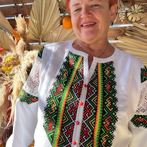 May include: A white embroidered blouse with a geometric pattern in red, green, orange, and black. The blouse has a V-neck and long sleeves with lace trim.