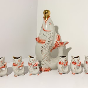 Op de afbeelding: Een witte porseleinen likeurset met een grote visvormige karaf met een gouden dop en zes kleinere visvormige shotglazen. De vissen zijn versierd met oranje en gouden details.