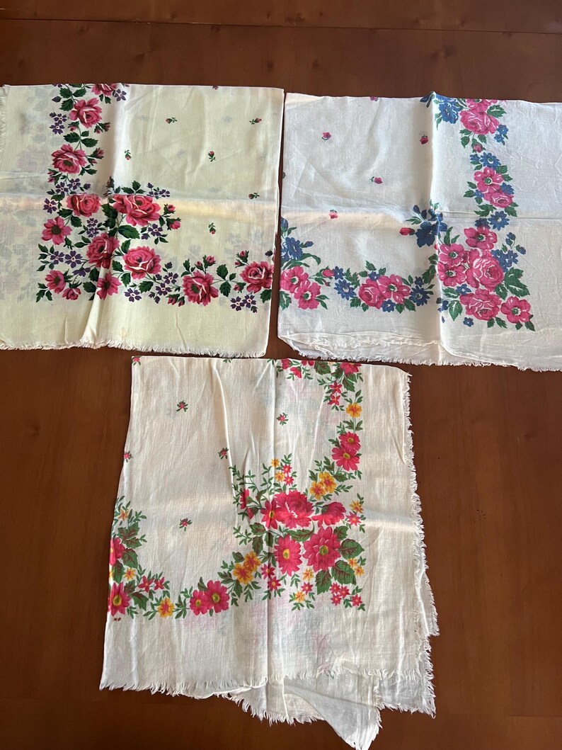 May include: Three white cotton handkerchiefs with floral patterns. The handkerchiefs have pink, yellow, blue, and green flowers. The handkerchiefs have a frayed edge.