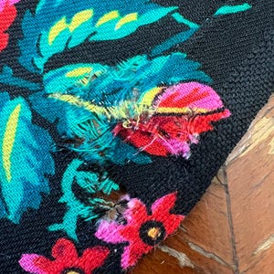 May include: Close-up of a black fabric with a floral pattern in shades of pink, red, yellow, and green. The fabric has a small tear in the upper right corner.