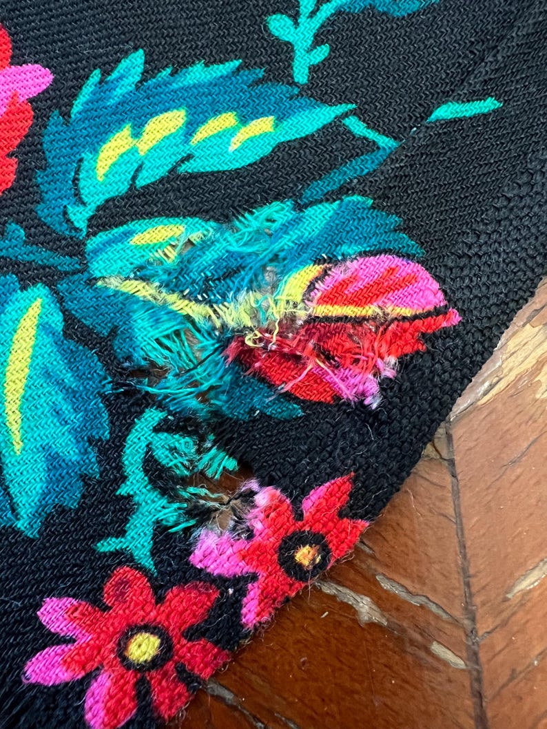 May include: Close-up of a black fabric with a floral pattern in shades of pink, red, yellow, and green. The fabric has a small tear in the upper right corner.