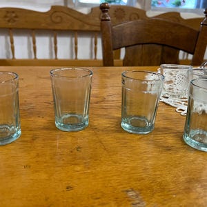 Puede incluir: Cuatro vasos pequeños y transparentes con un ligero tinte azulado están colocados sobre una mesa de madera. Los vasos son cilíndricos con una base gruesa. Un banco y una silla de madera están en el fondo.