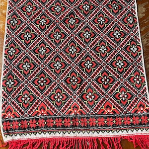 May include: A red, black and white embroidered fabric with a geometric pattern and a fringe border.