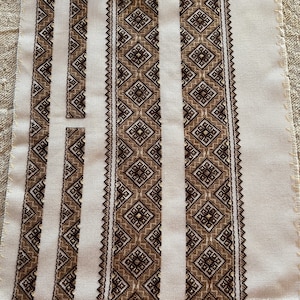Puede incluir: Un lienzo blanco con cinco tiras bordadas en marrón y negro. Las tiras presentan patrones geométricos.