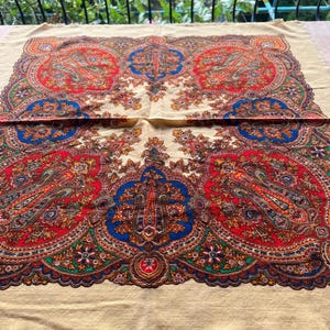 Puede incluir: Un chal beige con un vibrante estampado de cachemira en rojo, azul, naranja y verde. El intrincado diseño presenta motivos florales y de lágrima. El chal tiene un borde con flecos.