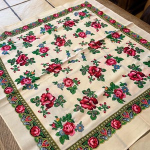 Puede incluir: Un pañuelo cuadrado de color blanco roto con un patrón repetitivo de rosas rojas vibrantes, hojas verdes y pequeñas flores azules. El pañuelo está enmarcado por un borde decorativo que contiene motivos de rosas rojas, follaje verde y formas azules.