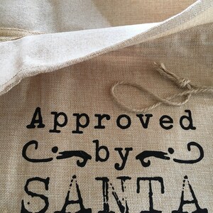 May include: A burlap gift bag with a brown string tie. The bag has black text that reads "Approved by Santa".