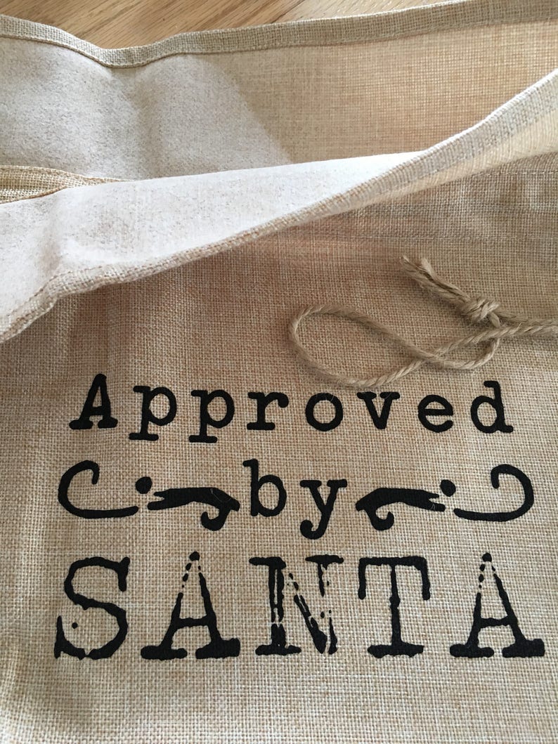 May include: A burlap gift bag with a brown string tie. The bag has black text that reads "Approved by Santa".
