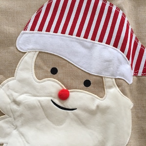 May include: A close-up of a Santa Claus face made of fabric. The face is white with a red nose and black eyes. The Santa Claus is wearing a red and white striped hat.
