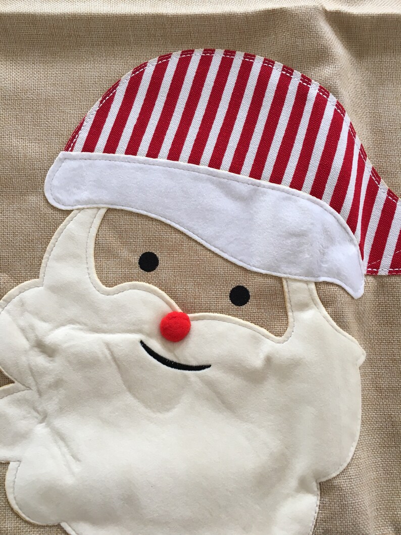 May include: A close-up of a Santa Claus face made of fabric. The face is white with a red nose and black eyes. The Santa Claus is wearing a red and white striped hat.