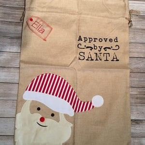May include: A brown burlap gift bag with a red and white striped Santa hat, a white beard, and the text "Approved by SANTA". A red tag with the name "Ella" is attached to the bag.