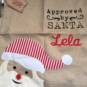 May include: A burlap Christmas stocking with a red and white striped Santa hat, a white beard, and a red nose. The stocking has the text "Approved by SANTA" and "Lela" embroidered on it.