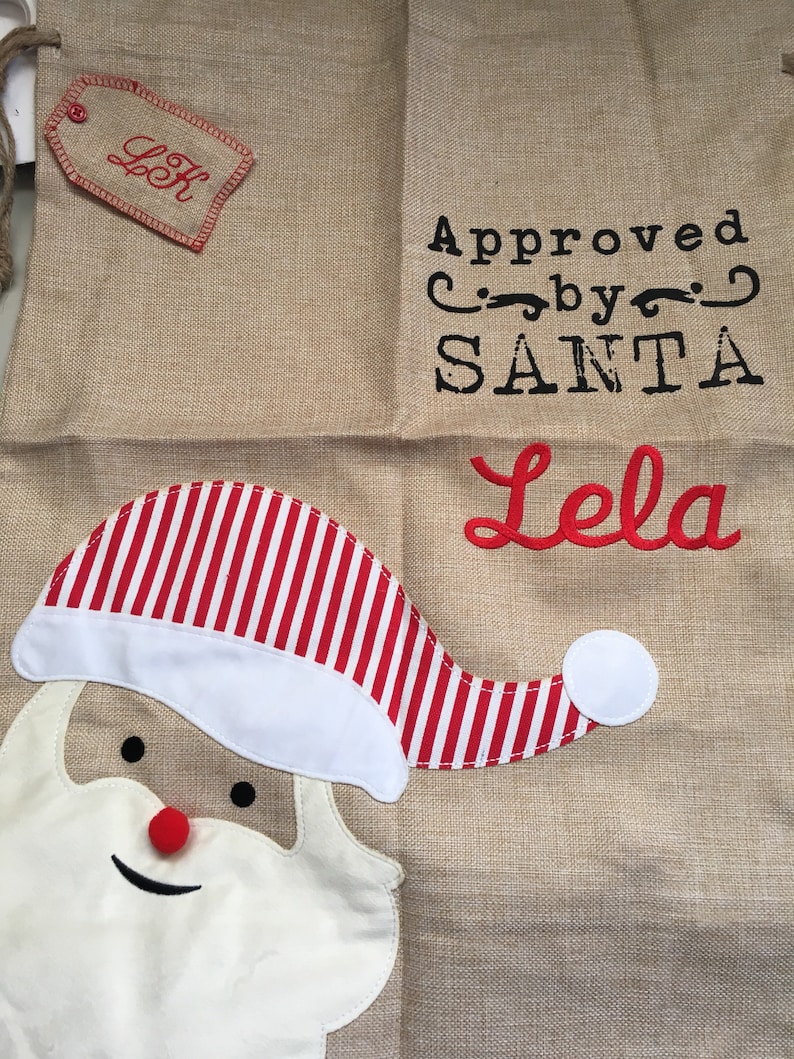 May include: A burlap Christmas stocking with a red and white striped Santa hat, a white beard, and a red nose. The stocking has the text "Approved by SANTA" and "Lela" embroidered on it.