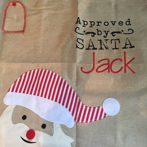 May include: A burlap sack with a Santa Claus face and the text "Approved by SANTA Jack". The sack has a red and white striped hat and a red tag.