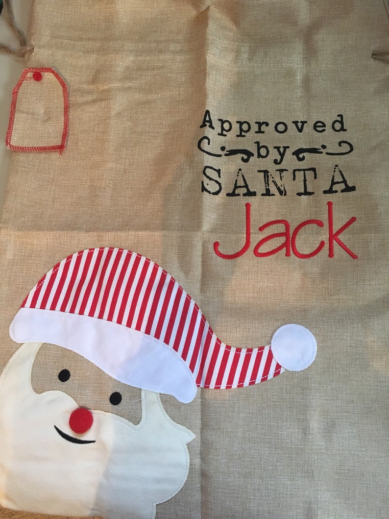 May include: A burlap sack with a Santa Claus face and the text "Approved by SANTA Jack". The sack has a red and white striped hat and a red tag.