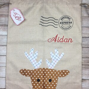 May include: A beige burlap Christmas gift bag with a red reindeer face, white polka dots, and red antlers. The bag has a red tag with the letter "B" on it and a black and white stamp that says "North Pole Express Reindeer Mail". The bag is personalized with the name "Aidan".