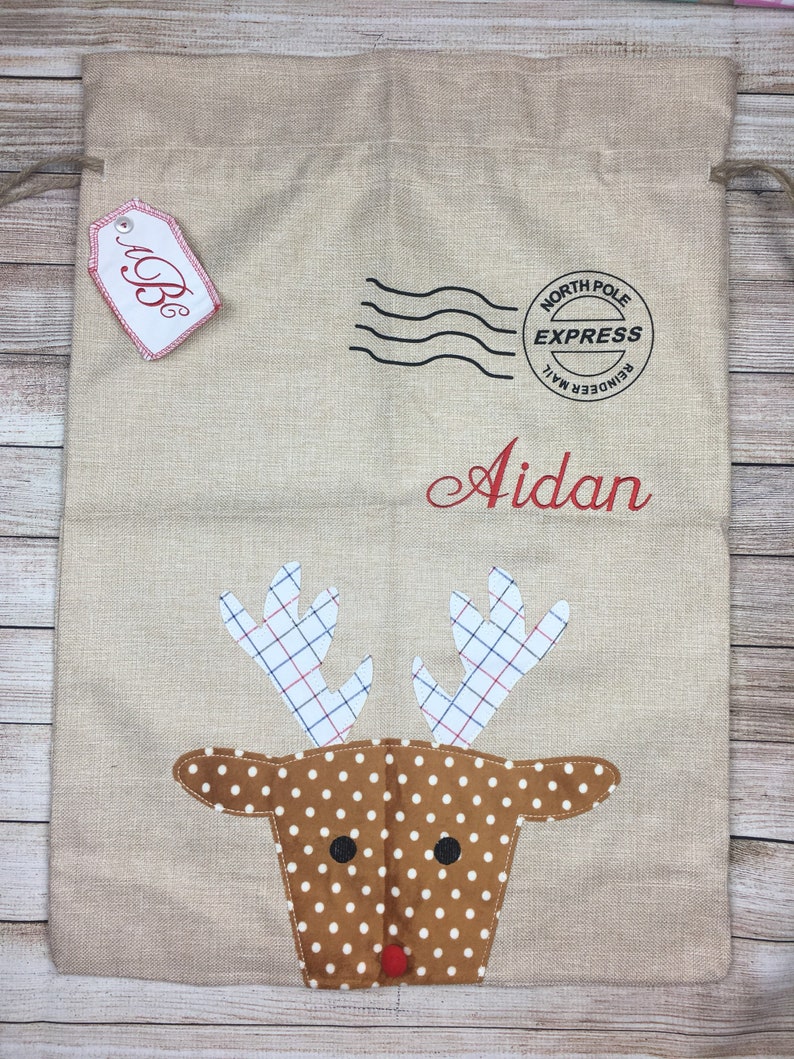 May include: A beige burlap Christmas gift bag with a red reindeer face, white polka dots, and red antlers. The bag has a red tag with the letter "B" on it and a black and white stamp that says "North Pole Express Reindeer Mail". The bag is personalized with the name "Aidan".