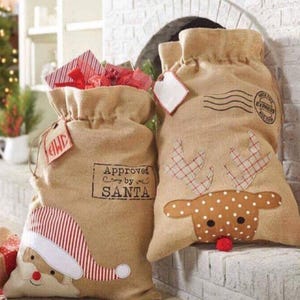 May include: Two burlap Christmas gift bags. One bag features a reindeer with red and white plaid antlers and brown polka dots. The other bag has a Santa Claus face with a red and white striped hat. Both bags have the text "Approved by Santa" printed on them.