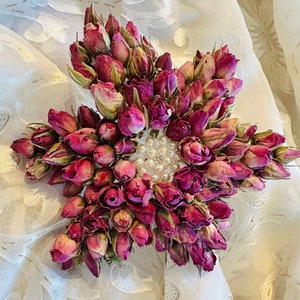 May include: A cluster of dried pink roses arranged in a star shape with a pearl-like center. The roses are surrounded by a white fabric background.