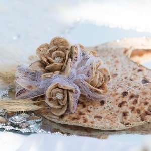 May include: A decorative arrangement of brown, woven flowers on a silver tray with a round, brown flatbread. The flowers are tied together with white tulle and have small white beads in the centre.