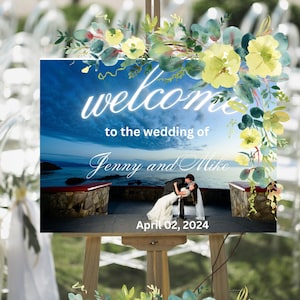 May include: A wedding welcome sign with a watercolor floral design in shades of blue and yellow. The sign features a photo of the couple against a scenic backdrop and the text "Welcome to the wedding of Jenny and Mike April 02, 2024".