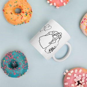 May include: White ceramic mug with a black line drawing of a bride and groom. The bride is wearing a wedding dress and the groom is wearing a suit. The drawing is surrounded by a heart and a string of flowers.