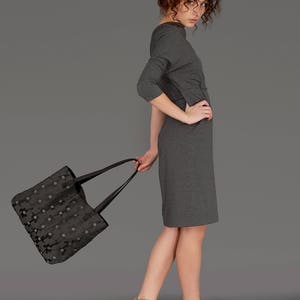 Op de afbeelding: Een vrouw in een grijze jurk houdt een zwarte tote bag met een bloemenpatroon vast. Ze draagt ​​bruine sandalen met hakken.