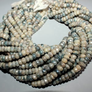 Puede incluir: Una hebra de cuentas de piedra preciosa facetadas de color azul claro y blanco. Las cuentas son redondas y tienen una forma ligeramente irregular. Las cuentas están enhebradas en un hilo blanco.