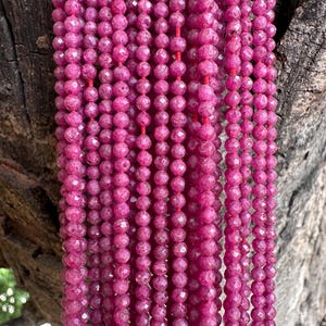 May include: Multiple strands of faceted, round, deep pink ruby beads are strung together on thin red thread. Each bead features a textured, reflective surface that catches the light, creating a uniform appearance across the vertical arrangement.