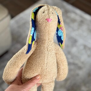 May include: A tan plush bunny with long ears. The inside of the ears are patterned with blue, green, and yellow circles on a navy background. The bunny has a pink nose and is being held by a person.