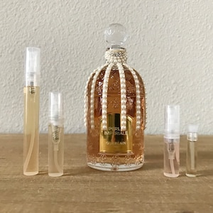 May include: A clear glass bottle of Guerlain perfume with a gold label that reads "Perle Imperiale". The bottle is decorated with white pearls and a gold cap. There are four smaller clear glass bottles of perfume next to the larger bottle.
