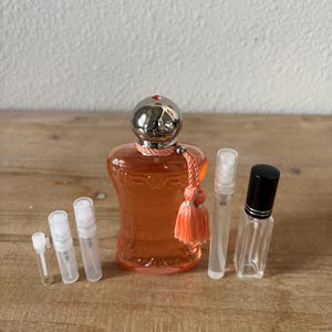 May include: An orange perfume bottle with a silver cap and a peach tassel, surrounded by several small, clear spray bottles. The perfume bottle is the focal point, with a decorative design. The bottles are arranged on a wooden surface.