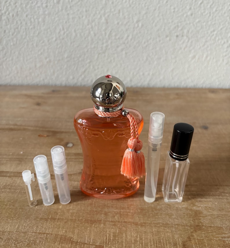 May include: An orange perfume bottle with a silver cap and a peach tassel, surrounded by several small, clear spray bottles. The perfume bottle is the focal point, with a decorative design. The bottles are arranged on a wooden surface.