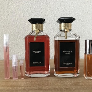 May include: Two large perfume bottles with black caps and gold accents, labeled "Patchouli Paris" and "Angelique Noire." Several smaller perfume vials are also present. The bottles are on a wooden surface.