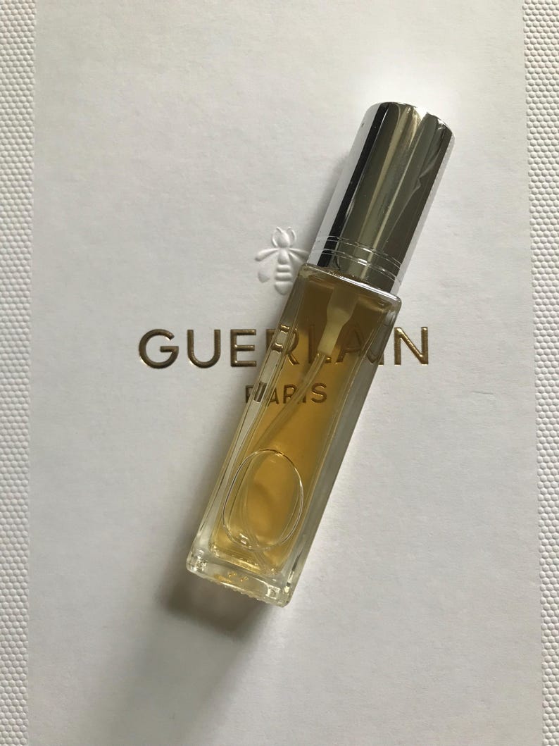 May include: A small, clear glass bottle of perfume with a silver cap. The bottle is filled with a light yellow liquid. The bottle is labelled "Guerlain Paris".