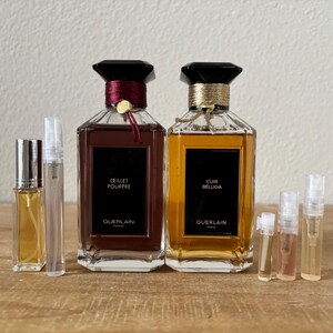 May include: Two Guerlain perfume bottles, one with a burgundy ribbon and the other with a gold ribbon, sit on a wooden surface. The bottles are labeled "Oeillet Pourpre" and "Cuir Beluga." Several small sample vials are also present.