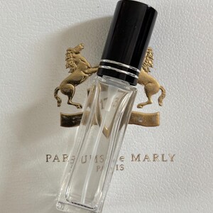 May include: A clear, rectangular perfume bottle with a black cap and silver accents. The bottle is angled, revealing its contents. The background features a white surface with a gold emblem and the text "PARFUMS de MARLY PARIS 1753."