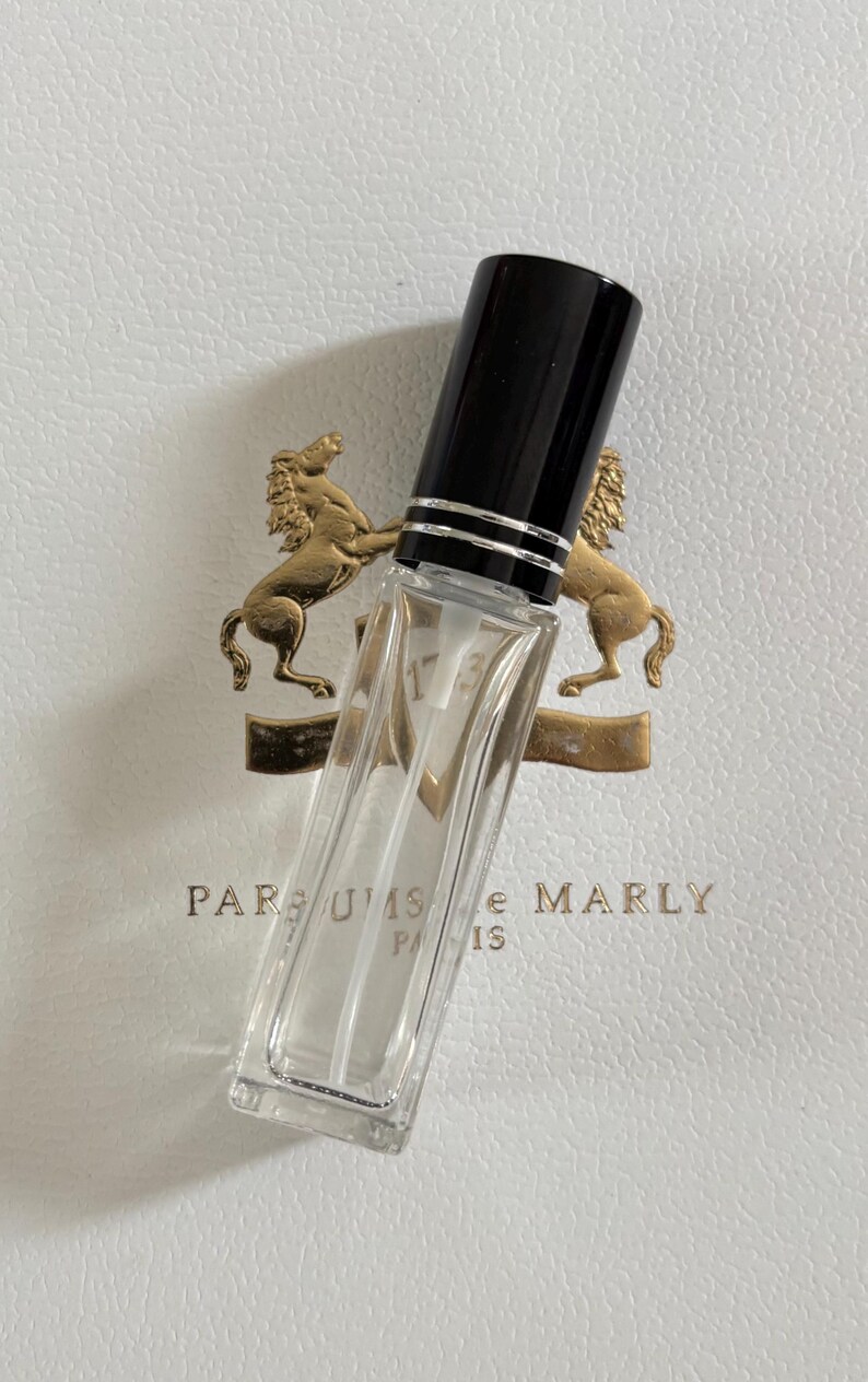 May include: A clear, rectangular perfume bottle with a black cap and silver accents. The bottle is angled, revealing its contents. The background features a white surface with a gold emblem and the text "PARFUMS de MARLY PARIS 1753."