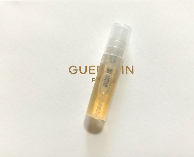 May include: A clear glass bottle containing a gold liquid. The bottle has a white cap and is labelled "Guerlain Paris".