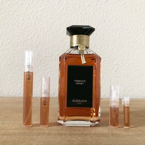 May include: A set of five perfume sample vials, four are clear and one is amber colored. The vials are arranged in a row on a wooden surface. A larger bottle of perfume is in the center of the image. The bottle is clear glass with a black cap and a gold band. The label on the bottle reads "Tobacco Honey" and "Guerlain Paris".