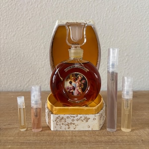 May include: A vintage perfume bottle with a round, amber-colored glass body and a decorative stopper. The bottle sits in a gold-colored box with a floral design. Several small perfume vials are also present, showcasing a fragrance collection.