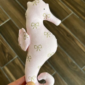 May include: A pink seahorse-shaped plush toy with gold bow patterns. The seahorse has a small eye and a curled tail. The toy is made of soft fabric and is designed for children.