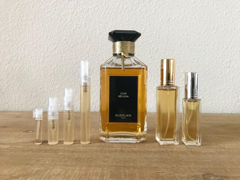 May include: A set of perfume bottles, including a large bottle of Guerlain Cuir Beluga perfume and several smaller sample bottles. The bottles are all clear glass with gold accents and contain a golden liquid.