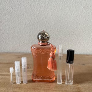 May include: An assortment of perfume bottles and sample vials. The main bottle is a peach-colored glass with a silver cap and a peach tassel. Several clear sample vials and a black-capped bottle are also present, all arranged on a wooden surface.