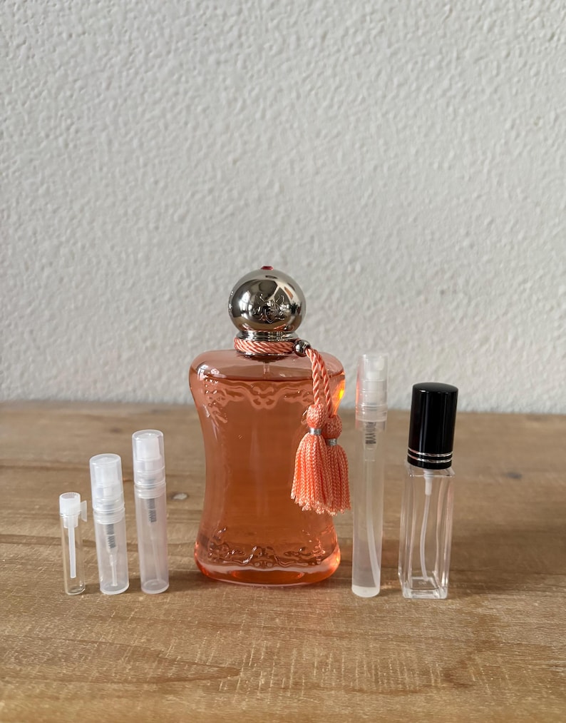 May include: An assortment of perfume bottles and sample vials. The main bottle is a peach-colored glass with a silver cap and a peach tassel. Several clear sample vials and a black-capped bottle are also present, all arranged on a wooden surface.